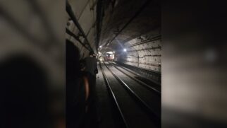 Fotografia em ângulo longitudinal tirada do interior de um túnel subterrâneo de metrô com pouca iluminação. Passageiros caminham em fila indiana pela estreita passarela lateral de emergência ao lado da via permanente e dos trilhos, enquanto a composição de um trem aparece paralisa ao fundo