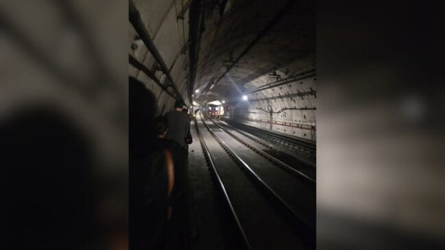 Fotografia em ângulo longitudinal tirada do interior de um túnel subterrâneo de metrô com pouca iluminação. Passageiros caminham em fila indiana pela estreita passarela lateral de emergência ao lado da via permanente e dos trilhos, enquanto a composição de um trem aparece paralisa ao fundo