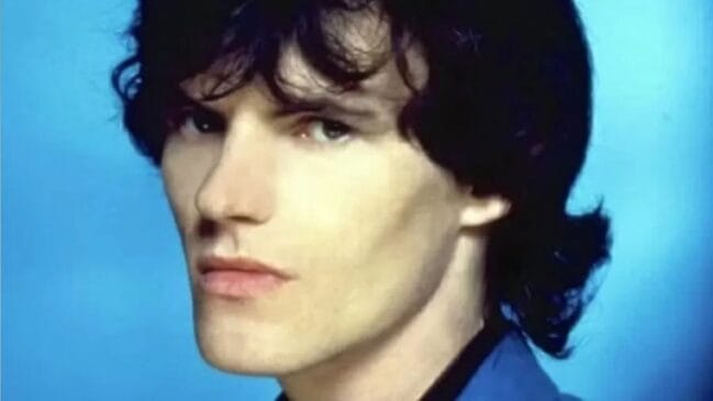 Retrato em plano fechado do cantor e músico Peter Byrne, vocalista da banda Naked Eyes, com cabelo escuro ondulado e a vestir um casaco azul sobre um fundo azul-claro