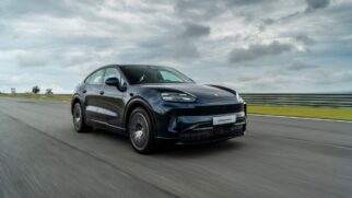 Fotografia em movimento de um SUV Porsche Cayenne azul-escuro a circular numa pista de testes de asfalto sob um céu nublado, captado em ângulo de três quartos dianteiro