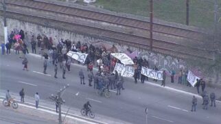 otografia aérea mostrando um grupo de manifestantes concentrado na berma de uma via junto a um muro com grafites e trilhos de trem paralelos ao fundo. Os protestantes seguram faixas brancas com as inscrições "S.O.S", "FAMÍLIAS EM RISCO", "JUSTIÇA" e "MORADIA". Agentes policiais e ciclistas acompanham a movimentação na pista