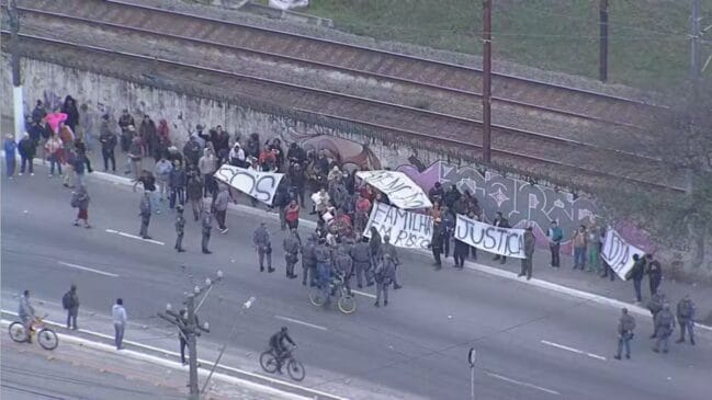 otografia aérea mostrando um grupo de manifestantes concentrado na berma de uma via junto a um muro com grafites e trilhos de trem paralelos ao fundo. Os protestantes seguram faixas brancas com as inscrições "S.O.S", "FAMÍLIAS EM RISCO", "JUSTIÇA" e "MORADIA". Agentes policiais e ciclistas acompanham a movimentação na pista