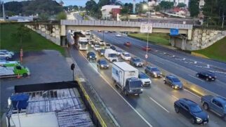 Fotografia de câmara de monitorização de tráfego numa autoestrada com fluxo intenso de veículos a passar sob um viaduto de betão. Em destaque, veem-se camiões, carrinhas e automóveis em circulação ao crepúsculo, com uma viatura de emergência estacionada na berma à esquerda