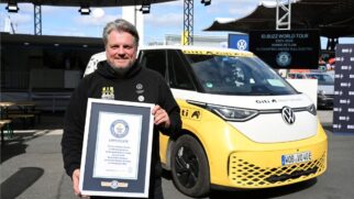 Fotografia em plano médio do piloto Rainer Zietlow a segurar um certificado emoldurado do Guinness World Records, posicionado à frente da carrinha elétrica Volkswagen ID.Buzz amarela e branca com adesivos de expedição