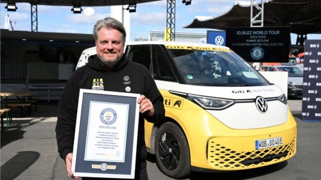 Fotografia em plano médio do piloto Rainer Zietlow a segurar um certificado emoldurado do Guinness World Records, posicionado à frente da carrinha elétrica Volkswagen ID.Buzz amarela e branca com adesivos de expedição