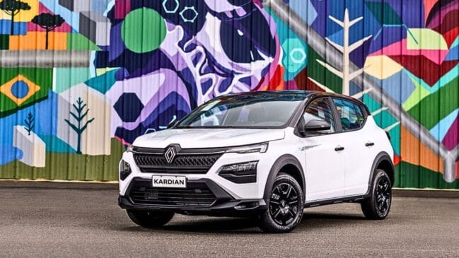 Fotografia diurna em ângulo três quartos dianteiro de um SUV compacto Renault Kardian branco com teto e retrovisores pretos. O veículo está estacionado diante de uma parede com painel de grafite colorido e geométrico. Destacam-se o logotipo da Renault centralizado na grade dianteira escura, os faróis afilados em LED divididos e as rodas de liga leve em tom preto