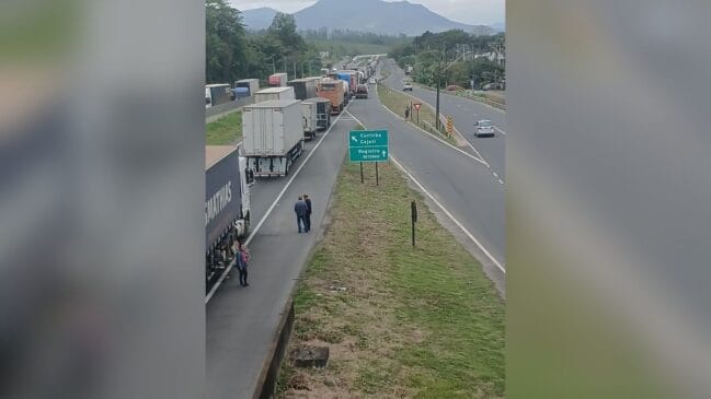 Fotografia em ângulo elevado de um longo congestionamento de caminhões e carretas retidos em uma rodovia. No canteiro central, destaca-se uma placa verde de sinalização viária com indicações para Curitiba, Cajati, Registro e Retorno. Motoristas e pedestres aparecem em pé no acostamento ao lado dos veículos parados, enquanto ao fundo observam-se serras e vegetação sob céu encoberto