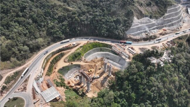 Fotografia aérea diurna em grande angular mostrando canteiro de obras de infraestrutura viária na Serra das Araras, na Rodovia Presidente Dutra (BR-116). Ao centro, observam-se pilares em construção para um novo viaduto, terraplanagem e contenções de encosta perfuradas no morro. Ao redor, a pista sinuosa da rodovia exibe o tráfego de caminhões e carros cercados pela vegetação densa de morros