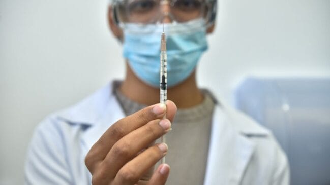 Fotografia em primeiro plano com foco centralizado em uma seringa graduada com dose de imunizante, segura pelas mãos de um profissional de saúde. Ao fundo, levemente desfocado, o profissional veste jaleco branco, máscara cirúrgica azul e óculos de proteção transparentes
