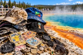 Cartas de Pokémon, chapéus, moedas e objetos retirados das águas termais de Yellowstone aparecem próximos a uma fonte azul cercada por vapor.