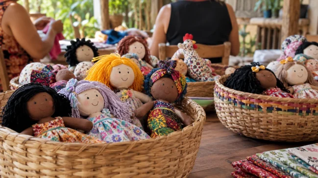 Representação de bonecos de pano de cores e formatos diferentes ficam reunidos em cestos de uma atividade comunitária. Imagem gerada por IA.