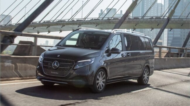 Fotografia de uma van executiva Mercedes-Benz Classe V (EQV) de cor cinzenta-escura em movimento sobre uma ponte estaiada, com perspetiva em ângulo de três quartos dianteiro, evidenciando a grelha frontal com o logótipo da estrela e detalhes luminosos em LED sob luz natural.