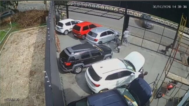 Imagem de câmara de segurança de um pátio ou concessionária de veículos mostrando vários automóveis estacionados, com indicação de data e hora no canto superior direito, e pessoas a afastar-se na sequência de um acidente ou impacto