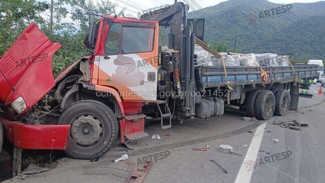 Caminhão vermelho envolvido em acidente rodoviário na berma de uma autoestrada, apresentando danos severos na cabina e na frente, com carga na carroçaria e encosta verde ao fundo, exibindo a marca d'água da Artesp