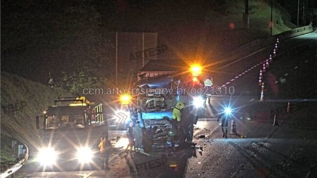 Fotografia noturna de um grave acidente rodoviário envolvendo um micro-ônibus com o para-brisas quebrado. Vários veículos de emergência com luzes laranja piscantes e equipes de socorro em coletes reflexivos estão no local. A pista está molhada e há destroços. Uma parede de contenção de concreto é visível ao fundo. A área está isolada com cones de trânsito