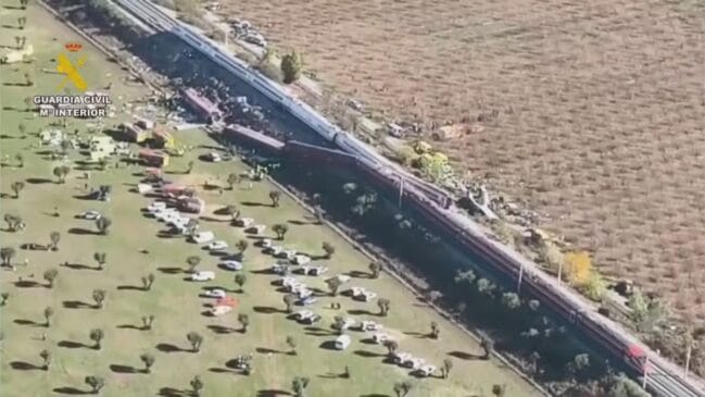 Vista aérea panorâmica de um descarrilamento ferroviário com carruagens de um comboio de passageiros saídas dos carris numa zona rural, exibindo o emblema oficial e texto da Guardia Civil e Ministério do Interior de Espanha no canto superior esquerdo