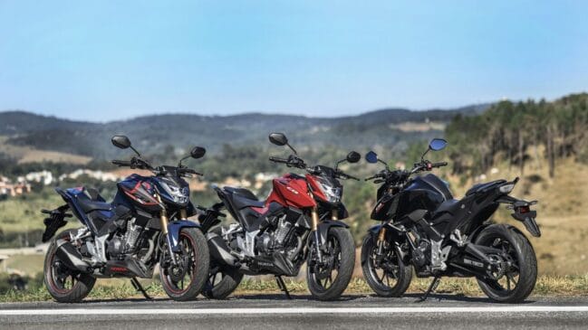 Três motocicletas Honda CB 300F Twister alinhadas lado a lado numa estrada ao ar livre, nas cores azul-escuro com detalhes vermelhos, vermelha e preta, com perspetiva em ângulo de três quartos dianteiro contra um fundo de montanhas e céu limpo.