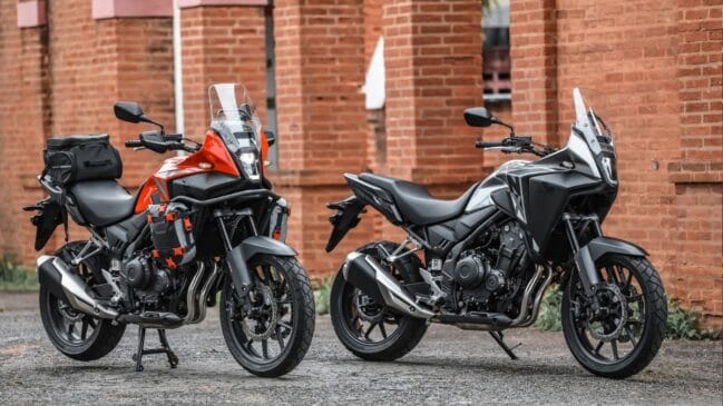 Fotografia de duas motocicletas crossover Honda NX500 estacionadas lado a lado em frente a uma parede de tijolo vermelho, sendo o modelo à esquerda em tom laranja com acessórios de viagem e malas, e o modelo à direita em cor escura