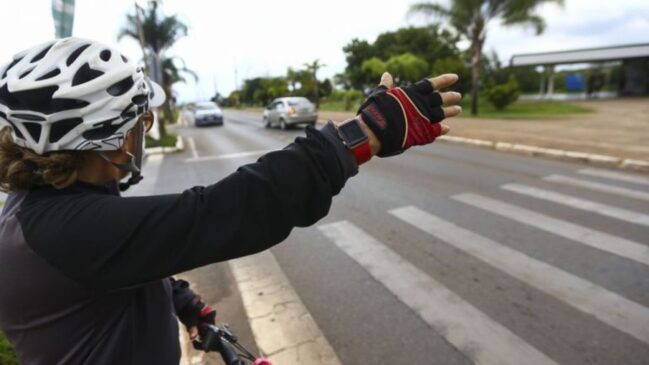 Ciclista equipada com capacete branco e luvas de ciclismo parada junto a uma passadeira numa rua asfaltada, estendendo o braço para sinalizar a travessia, com automóveis em circulação ao fundo