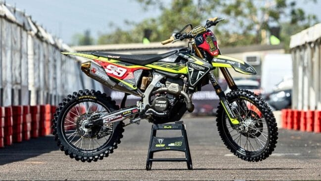 Motocicleta de motocross Triumph TF250-X com grafismo de competição nas cores preto e amarelo, equipada com número de corrida, escape desportivo e pneus de cravos, apoiada num cavalete de paddock ao ar livre