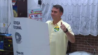 Fotografia do político Flávio Bolsonaro a votar numa cabina de voto da Justiça Eleitoral no Rio de Janeiro, vestindo uma camisa amarela e a fazer um gesto positivo com o polegar