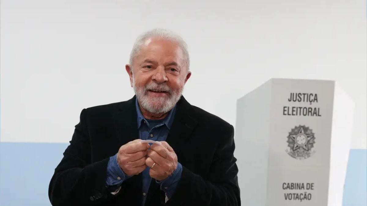 O Presidente da República, Luiz Inácio Lula da Silva, sorridente em plano médio, segura o comprovativo de votação em papel com ambas as mãos, estando posicionado ao lado de uma cabina de votação de papelão da Justiça Eleitoral