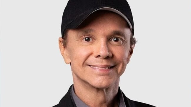Retrato de rosto e ombros do cantor Netinho (Netinho da Bahia, ex-É o Tchan) sorrindo, vestindo um boné de beisebol preto e uma jaqueta preta sobre uma camisa cinza, contra um fundo cinza neutro desfocado