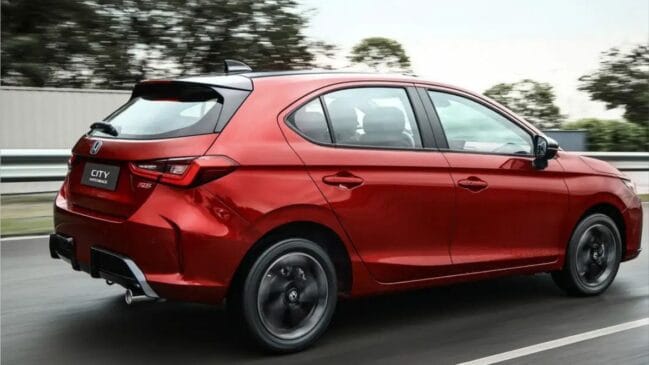 Fotografia traseira de um Honda City Hatchback vermelho em movimento num dia nublado. O carro exibe jantes de liga leve escuras e o emblema "CITY HATCHBACK" na tampa da mala, com a paisagem da estrada desfocada ao fundo