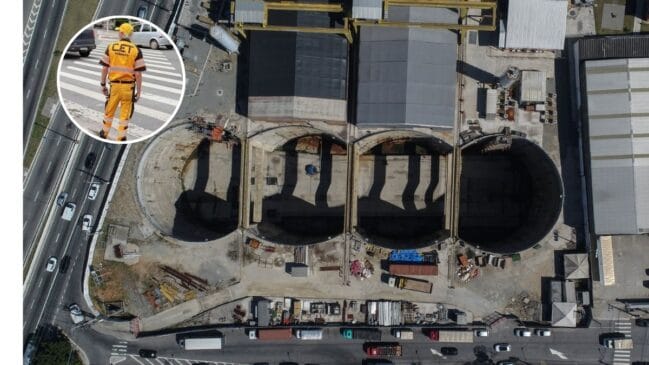 Vista aérea do estaleiro de obras da Linha 6-Laranja do Metro de São Paulo, mostrando grandes poços circulares de escavação e infraestruturas operacionais ao lado de uma via com tráfego. No canto superior esquerdo, um detalhe circular em destaque exibe um agente da Companhia de Engenharia de Tráfego (CET) de costas, com uniforme amarelo e preto, numa passagem de peões