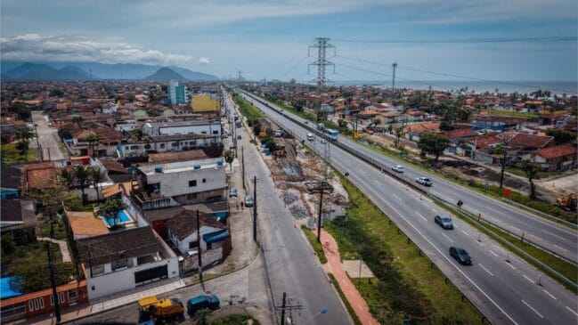 Vista aérea panorâmica de uma cidade litorânea cruzada por uma rodovia duplicada e uma via marginal com obras, mostrando torres de transmissão de energia, ciclovia vermelha, casas e o oceano ao fundo sob céu ensolarado