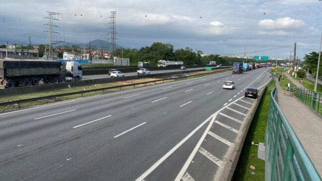 Vista panorâmica de uma rodovia movimentada em Cubatão, com múltiplas faixas de rodagem onde circulam automóveis e camiões pesados, ladeada por um passeio com guarda-corpo verde à direita, torres de alta tensão e vegetação ao fundo sob céu nublado