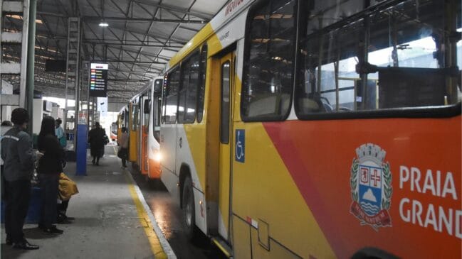 Autocarro urbano amarelo e branco do transporte municipal de Praia Grande estacionado na plataforma de um terminal coberto, exibindo o brasão do município e o símbolo internacional de acessibilidade na porta lateral, com passageiros a aguardar no cais à esquerda