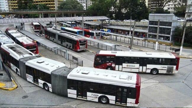 Vista aérea e panorâmica de um grande terminal de autocarros urbano em São Paulo, Brasil, com múltiplos autocarros articulados e standard a circular e estacionados nas plataformas. A infraestrutura inclui um corredor de autocarros elevado e curvo, passarelas para peões e edifícios urbanos no fundo. Autocarros com a inscrição "7 6329" são visíveis em primeiro plano