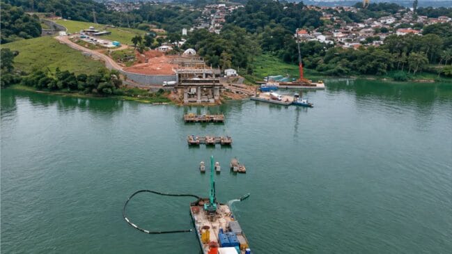 Vista aérea das obras de construção da ponte do Grajaú sobre uma represa, mostrando pilares em betão, equipamentos de engenharia e barcaças na água, com margens arborizadas ao fundo
