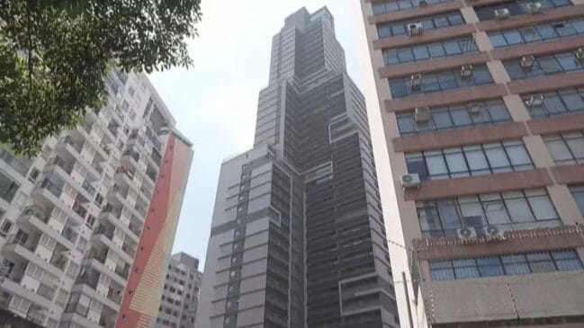 Perspetiva contra-plongeante de um edifício alto escuro no centro urbano, ladeado por outros prédios comerciais e residenciais com janelas e varandas