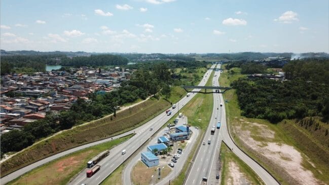 Vista aérea panorâmica do Rodoanel Mário Covas, operado pela SPMAR, com múltiplos veículos a circular nas faixas de rodagem, uma praça de apoio com edifícios de telhado azul, áreas residenciais urbanas e vegetação circundante sob um céu com nuvens dispersas