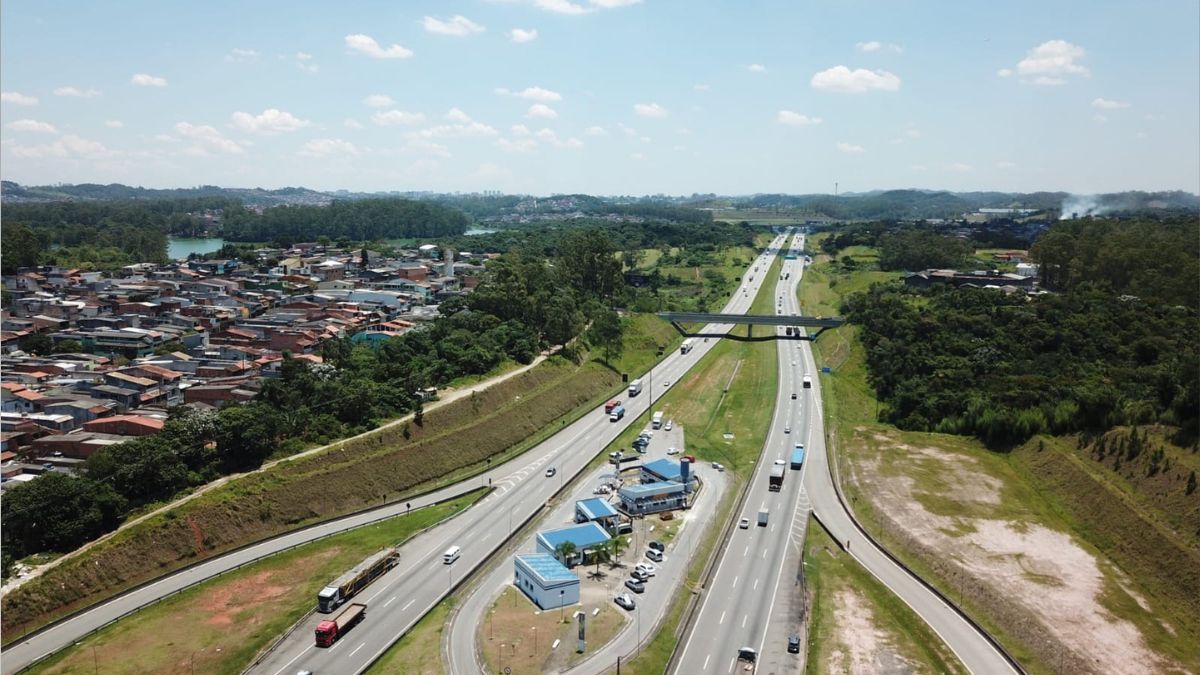 Vista aérea panorâmica do Rodoanel Mário Covas, operado pela SPMAR, com múltiplos veículos a circular nas faixas de rodagem, uma praça de apoio com edifícios de telhado azul, áreas residenciais urbanas e vegetação circundante sob um céu com nuvens dispersas