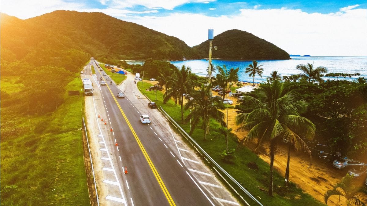 Vista aérea da Rodovia Rio-Santos (BR-101) contornando a costa litorânea, com veículos a circular nas faixas, vegetação de Mata Atlântica nas encostas, palmeiras e a praia com o mar ao fundo num dia ensolarado
