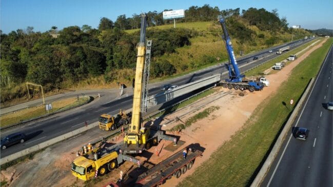 Vista aérea de obras de infraestrutura numa rodovia, onde duas grandes gruas telescópicas (uma amarela e uma azul) posicionam uma viga pré-moldada de betão/concreto sobre o canteiro lateral, enquanto o tráfego de veículos flui nas faixas de rodagem ao lado