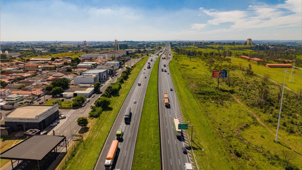 Vista aérea panorâmica da Rodovia dos Bandeirantes, com múltiplos veículos e camiões a circular nas faixas de rolamento, rodeada por áreas verdes e infraestruturas urbanas e industriais
