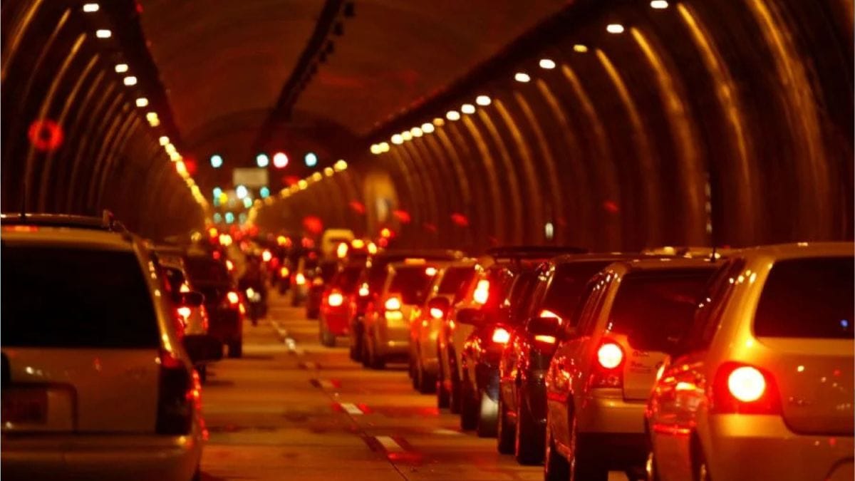 Perspetiva interna de um túnel urbano com tráfego automóvel intenso e lento, mostrando as luzes traseiras vermelhas dos veículos acesas e a iluminação amarelada do teto refletida ao longo das vias