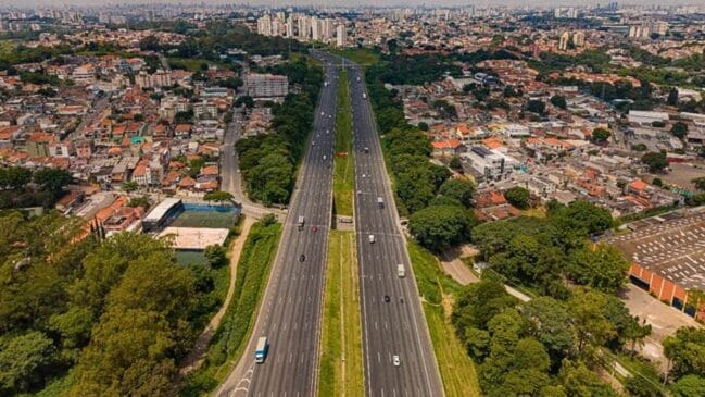 Vista aérea em perspetiva de uma autoestrada de múltiplas faixas duplicadas (Sistema Anhanguera-Bandeirantes) a cortar uma malha urbana densa, com tráfego fluido de veículos e canteiro central relvado sob luz solar