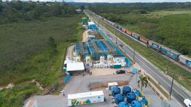 Vista aérea panorâmica de uma estação de tratamento de água e efluentes da Sabesp em área verde, mostrando tanques circulares azuis, reservatórios e módulos operacionais paralelos a uma rodovia e a uma linha férrea por onde passa um comboio de mercadorias da Rumo
