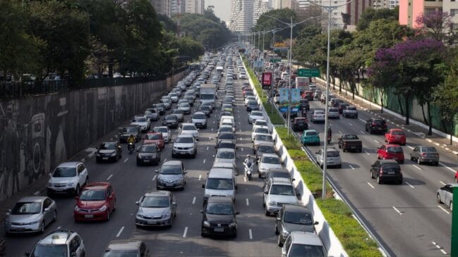 Vista elevada e em perspetiva do tráfego intenso na Avenida 23 de Maio, em São Paulo, com múltiplas faixas de rodagem marcadas por longas filas de carros e veículos comerciais engarrafados em direção ao centro da cidade, ladeadas por árvores e edifícios ao fundo