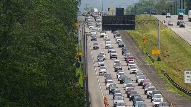 Perspetiva elevada de uma autoestrada com tráfego intenso e engarrafamento de veículos ao longo das faixas de rodagem, com vegetação densa à esquerda e painéis de mensagem variável da Polícia Rodoviária e concessionária ao longo da pista